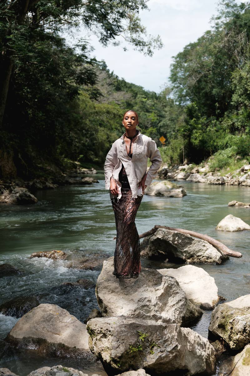 Model Christabelle Brown radiates quiet strength and mystery in an asymmetrical stone grey linen long-sleeve shirt with raw hems and textured details that mirror the rugged river landscape. Beneath, a flowing black lace dress adds softness and movement aga