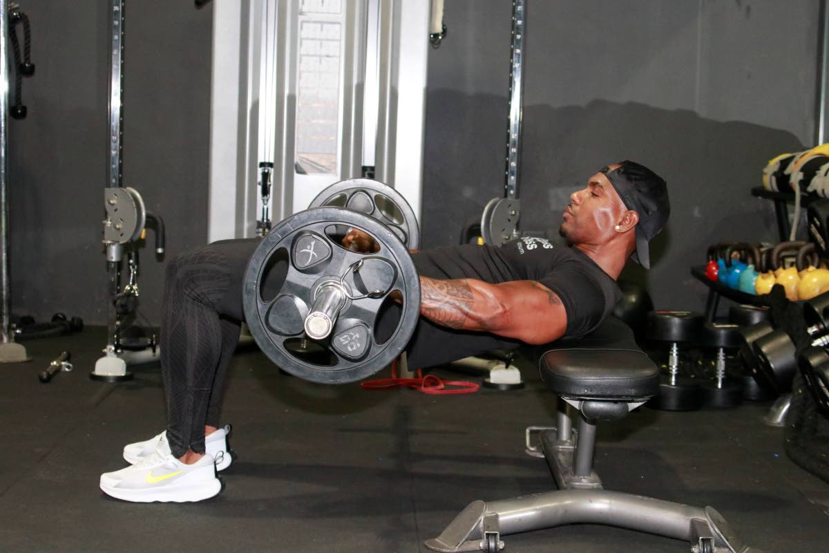 Brown demonstrates the hip thrust exercise, with shoulders on a bench and weight placed on the hips, lifting the hips upward in a controlled motion.