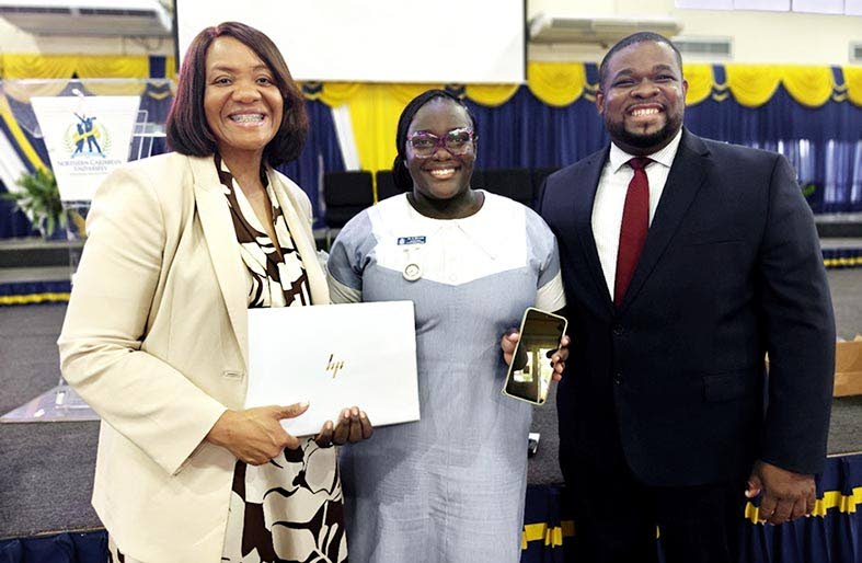 Anastacia Williams (centre) shows the mobile phone she relied on for university coursework alongside the laptop she received through a partnership involving AdventHealth and Andrews Memorial Hospital. Sharing in the moment are Dr Audrey Gregory (left), sen