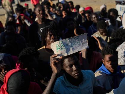 A migrant who crossed from Morocco into Spain holds a sign reading, in Spanish, “asylum” as he lines up for food distribution on a beach in the Spanish enclave of Ceuta, Monday, August 3, 2026. 