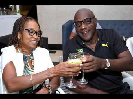 Lois Spence, director of business development and marketing at Covenant Insurance Brokers, clinks glasses with a smiling Bertland Cameron, veteran track and field coach and Olympian, as they watch the Paris Olympics opening ceremony from the comfort of The